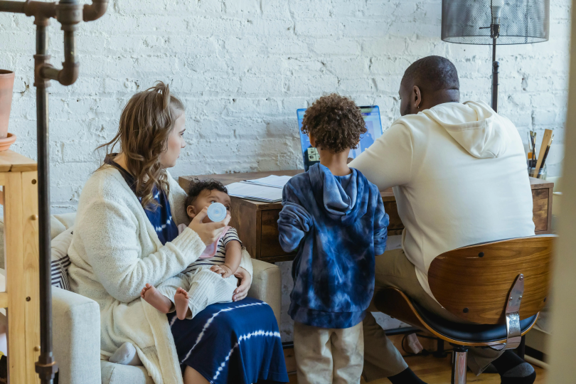 Family of four sitting around the table Father and child are in front of the computer and mother is feeding the baby.